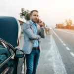 A man in a gray blazer and blue shirt stands beside a black car with its hood open, talking on a phone near the roadside at sunset.