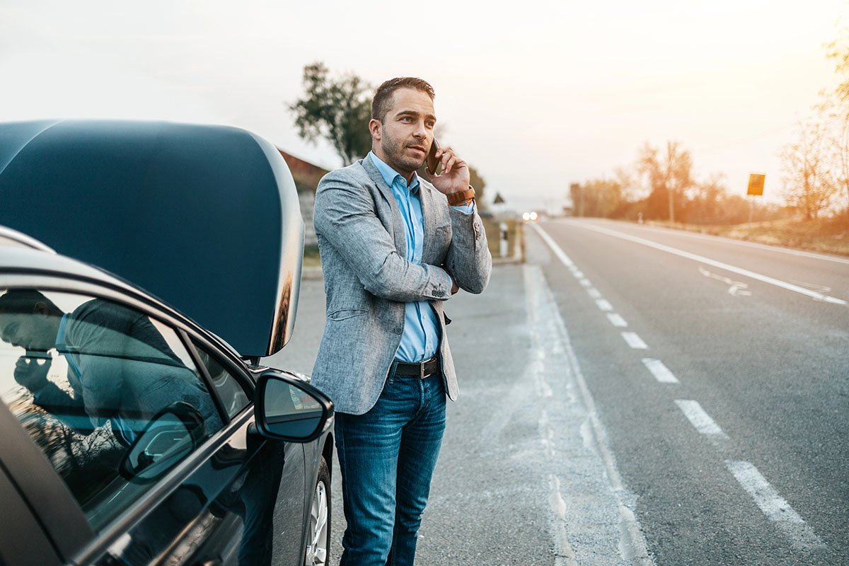 A man in a gray blazer and blue shirt stands beside a black car with its hood open, talking on a phone near the roadside at sunset.