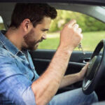 A frustrated man grips a steering wheel inside a car, holding car keys and grimacing as he focuses on the dashboard.