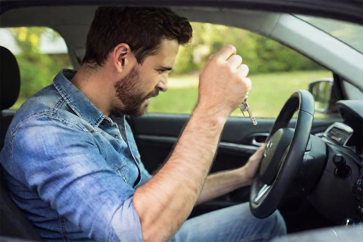 A frustrated man grips a steering wheel inside a car, holding car keys and grimacing as he focuses on the dashboard.