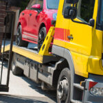 A yellow tow truck is in the process of loading a red car onto its flatbed, with a brick pillar and gate partially visible in the foreground.