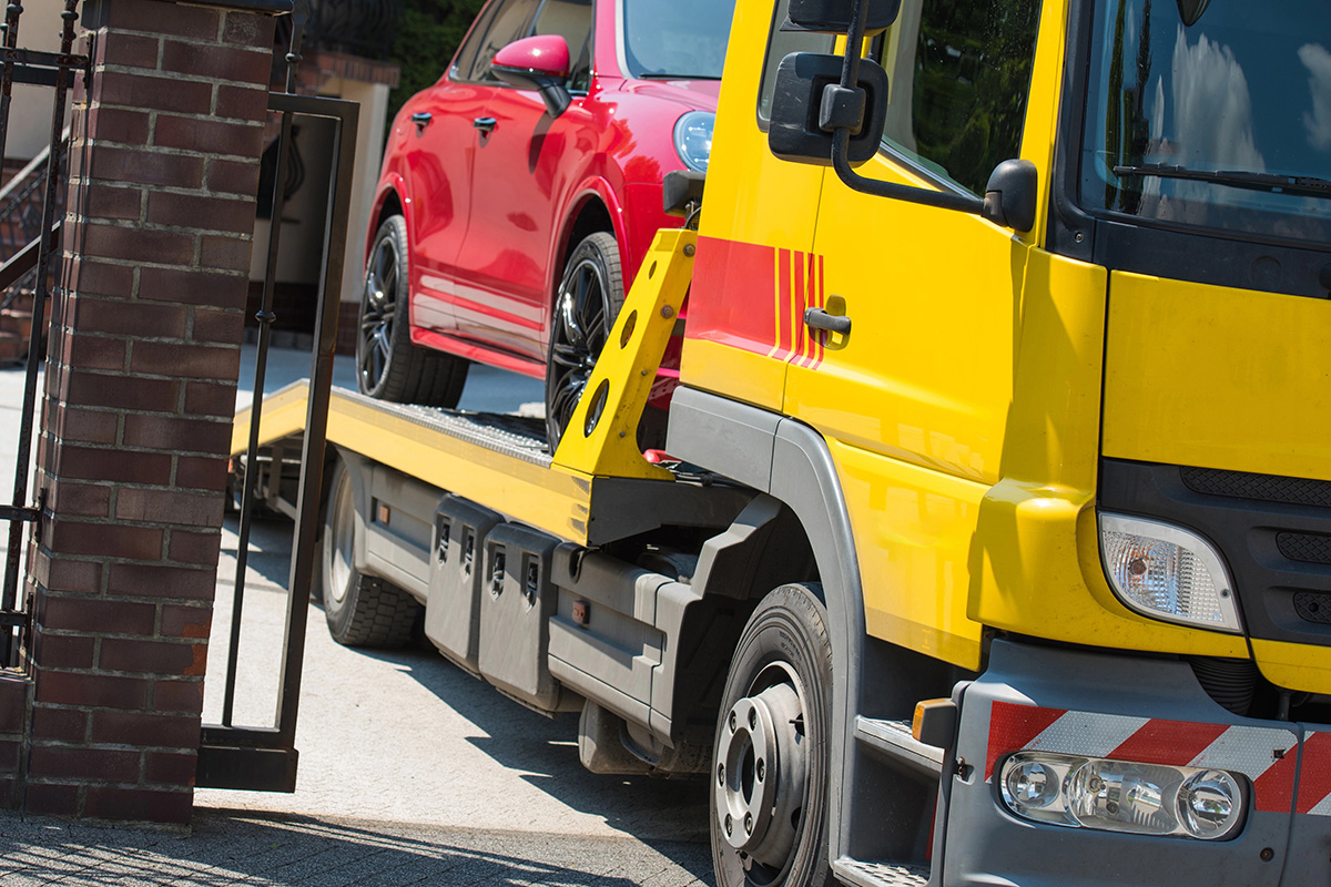 A yellow tow truck is in the process of loading a red car onto its flatbed, with a brick pillar and gate partially visible in the foreground.