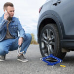 A young man in a denim jacket squats beside a car with a flat tire, talking on his phone while holding a wrench. A blue car jack and a tire iron are on the ground nearby.
