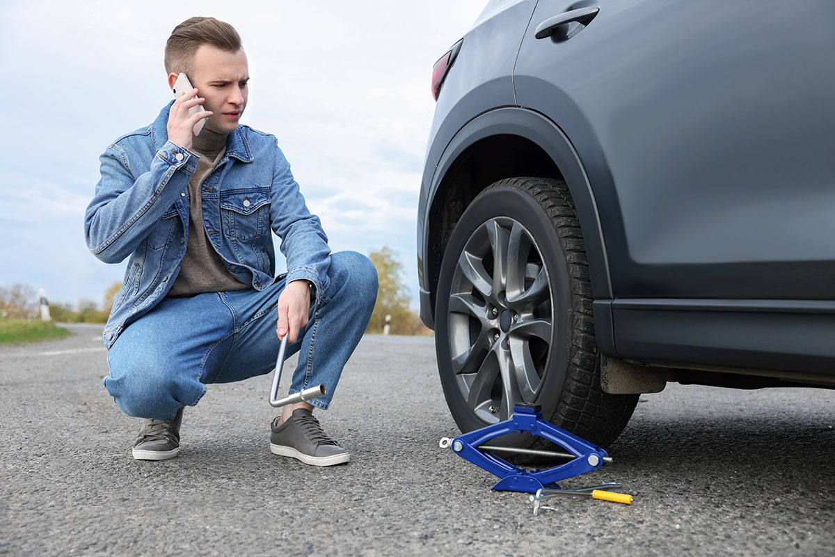 A young man in a denim jacket squats beside a car with a flat tire, talking on his phone while holding a wrench. A blue car jack and a tire iron are on the ground nearby.