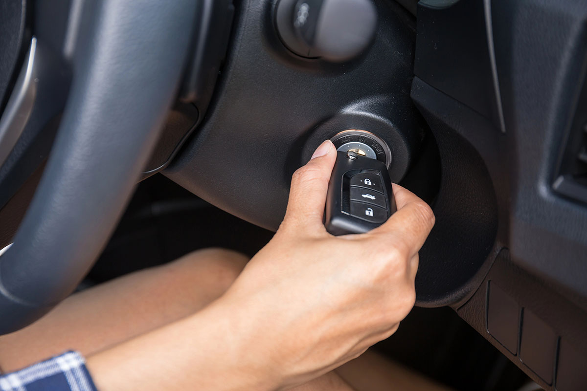 A hand is inserting a car key into the ignition of a vehicle, with the steering wheel and part of the dashboard visible in the background.