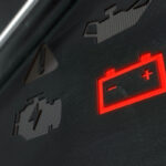 A close-up of a vehicle dashboard displaying a glowing red battery warning icon, indicating a potential issue with the battery. Other warning symbols are faintly visible in the background.