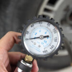 A person holds a pressure gauge displaying readings in PSI and BAR, with a blurred tire in the background.