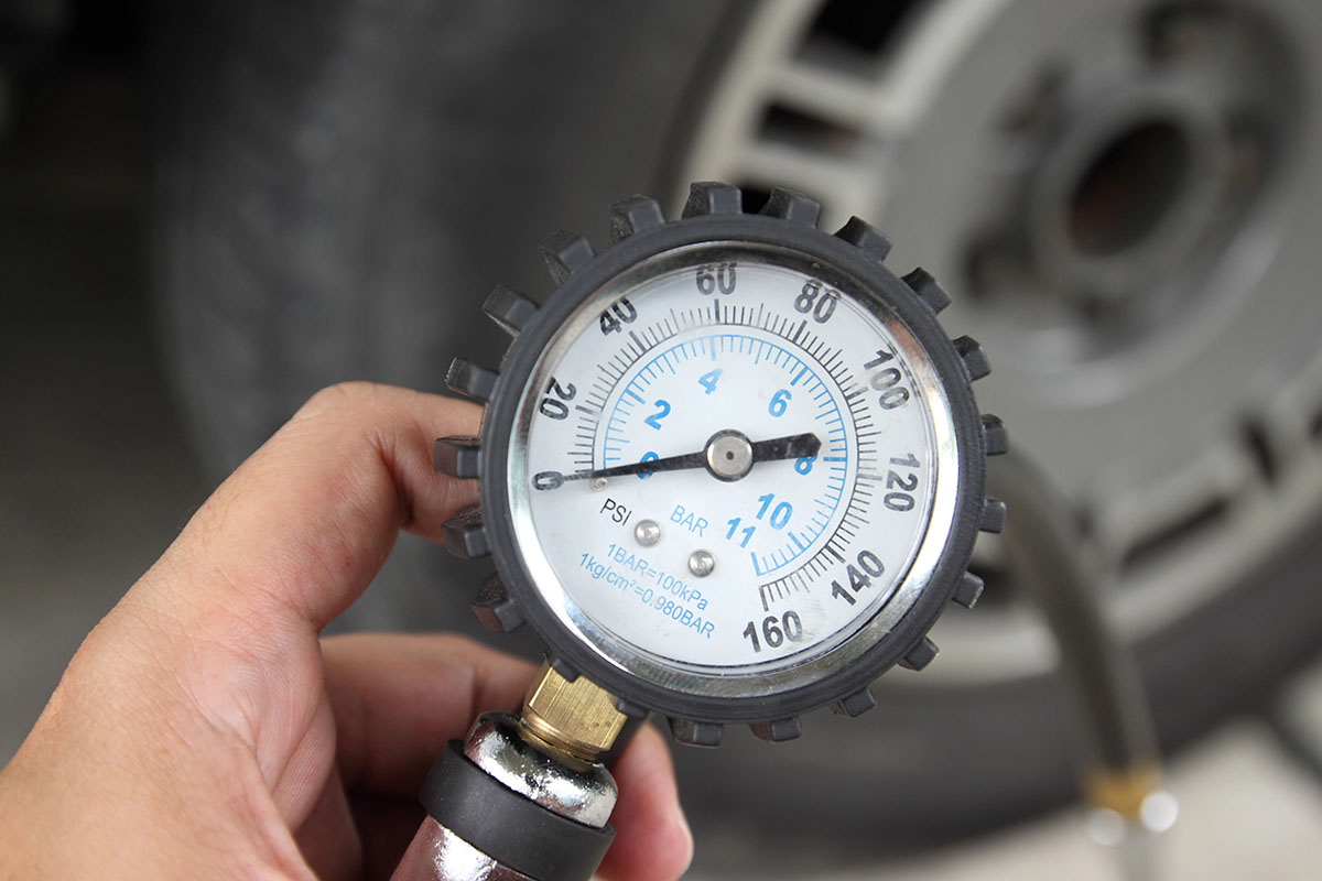 A person holds a pressure gauge displaying readings in PSI and BAR, with a blurred tire in the background.