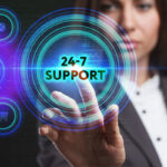A business professional in a suit points toward a digital button labeled "24-7 SUPPORT," surrounded by various icons, including an envelope, globe, and shopping cart, against a dark, tech-themed background.