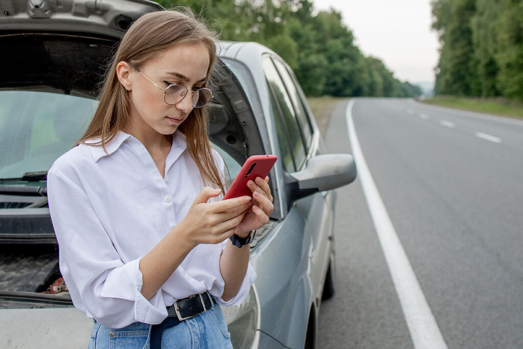 A woman wearing glasses and a white shirt stands beside a car with its hood open, looking at her phone while parked on the side of a road surrounded by trees.