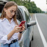 A woman wearing glasses and a white shirt stands beside a car with its hood open, looking at her phone while parked on the side of a road surrounded by trees.