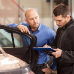 Two men converse in a car repair shop; one, in a blue shirt, leans against a car door, while the other, wearing a black mechanic's jumpsuit and gloves, takes notes on a clipboard.