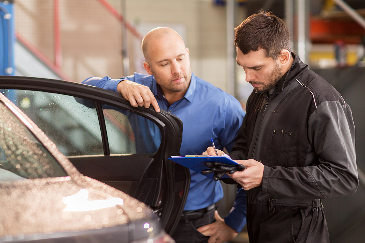 Two men converse in a car repair shop; one, in a blue shirt, leans against a car door, while the other, wearing a black mechanic's jumpsuit and gloves, takes notes on a clipboard.