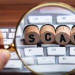 A hand holding a magnifying glass over a computer keyboard, focusing on wooden blocks that spell "SCAM."