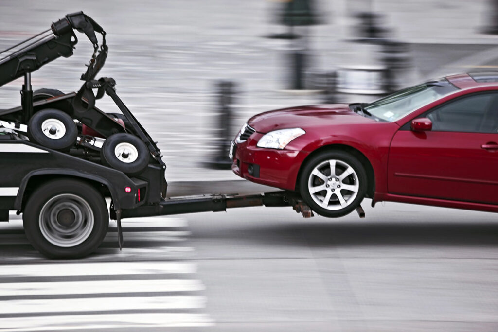 A red car is being towed by a tow truck, with the towing mechanism partially raised, moving across a crosswalk.