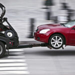 A red car is being towed by a tow truck, with the towing mechanism partially raised, moving across a crosswalk.