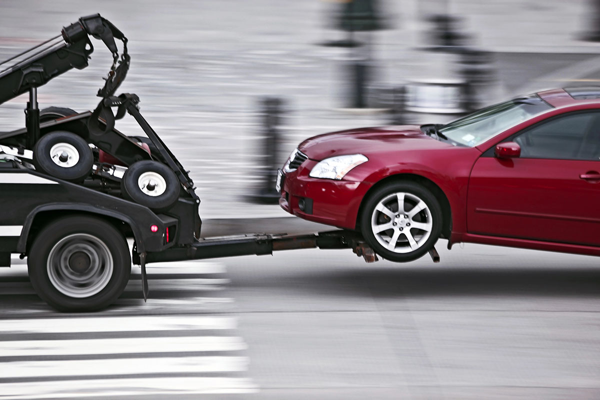 A red car is being towed by a tow truck, with the towing mechanism partially raised, moving across a crosswalk.