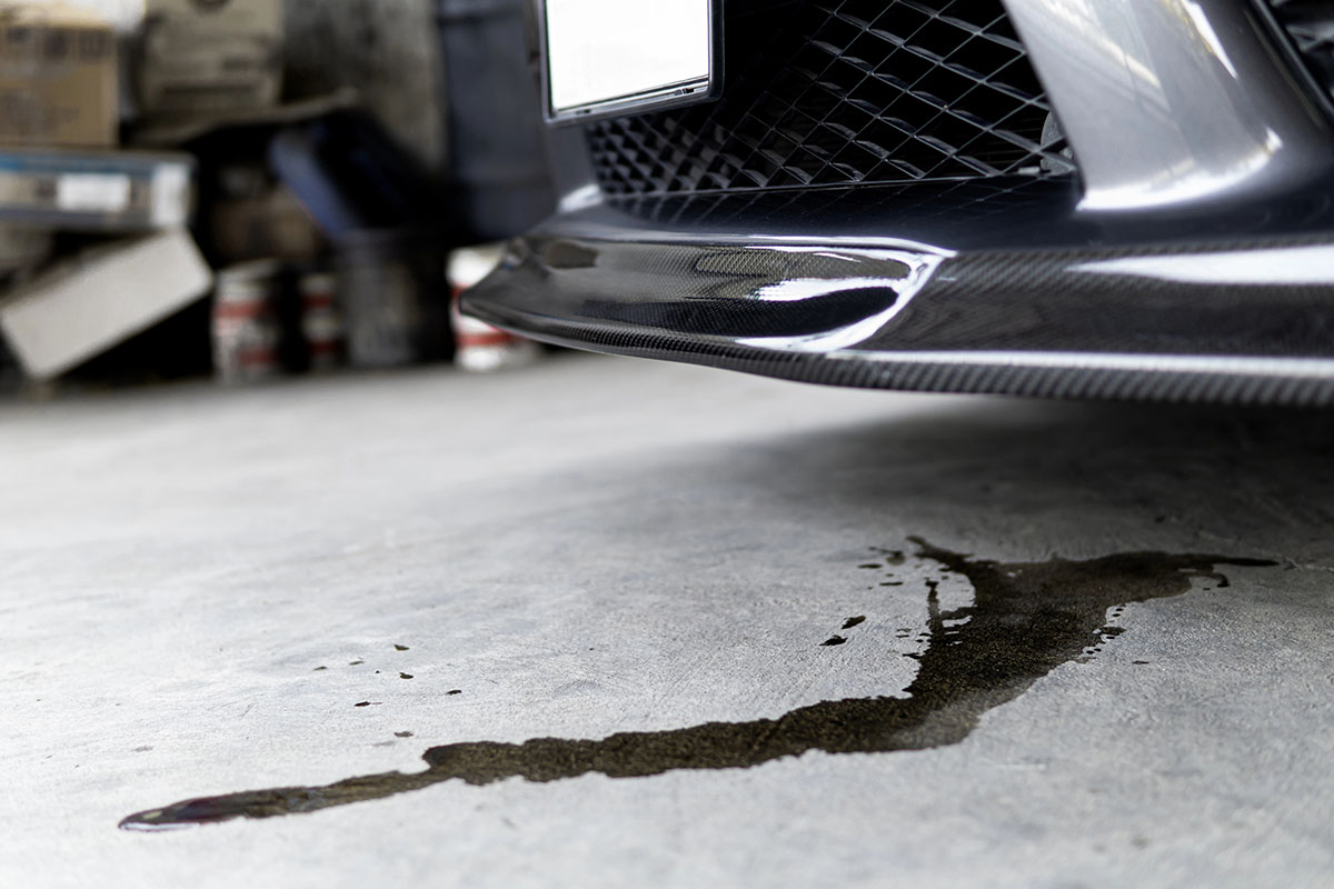 Close-up of a car's front bumper with a puddle of brownish fluid on the concrete floor beneath it, suggesting a potential leak. Background features boxes and paint cans.