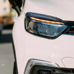 Close-up of a modern vehicle's headlight, featuring a sleek design with a prominent LED light strip and a reflective lens, set against a blurred background of another car and urban scenery.