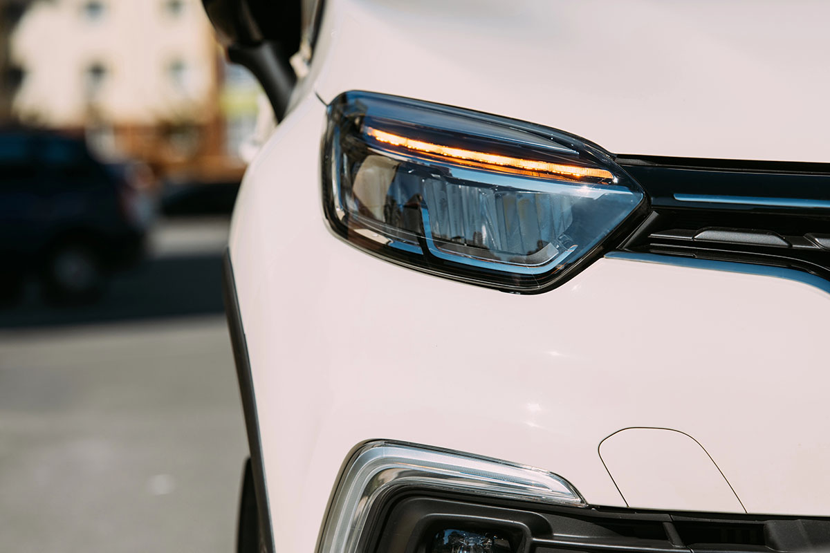 Close-up of a modern vehicle's headlight, featuring a sleek design with a prominent LED light strip and a reflective lens, set against a blurred background of another car and urban scenery.