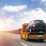 A tow truck carrying a black car on its flatbed drives on a highway with a clear sky and soft clouds in the background.