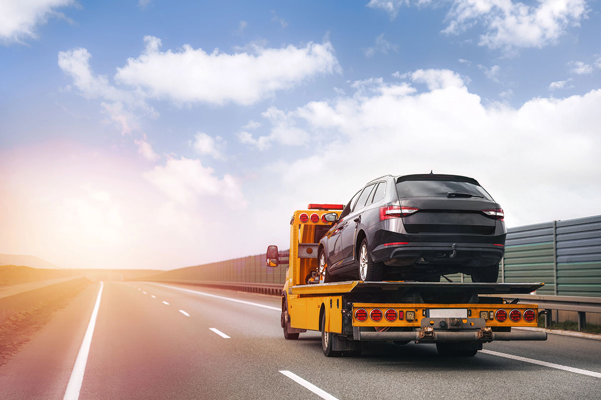 A tow truck carrying a black car on its flatbed drives on a highway with a clear sky and soft clouds in the background.