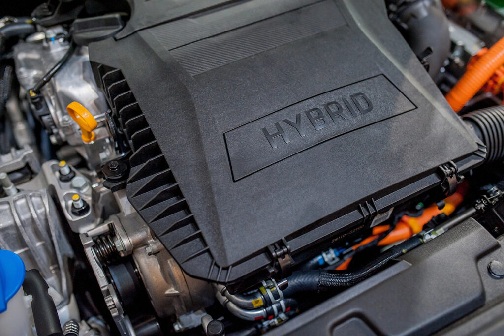 Close-up of a hybrid engine, featuring a prominent plastic cover labeled "HYBRID," with visible electrical components, hoses, and a bright orange wire, highlighting the engine's modern technology.