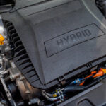 Close-up of a hybrid engine, featuring a prominent plastic cover labeled "HYBRID," with visible electrical components, hoses, and a bright orange wire, highlighting the engine's modern technology.