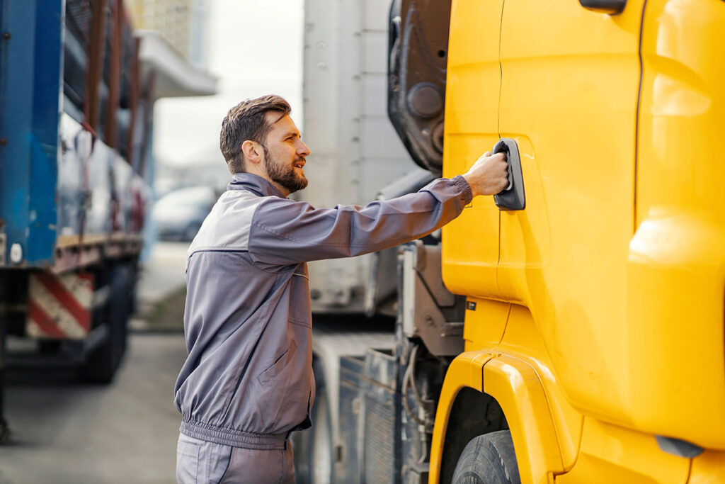 A man in a gray work uniform is reaching for the handle on the door of a yellow truck, standing next to another parked truck in an urban setting.