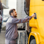 A man in a gray work uniform is reaching for the handle on the door of a yellow truck, standing next to another parked truck in an urban setting.