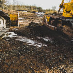 Two heavy machinery vehicles are parked on a muddy construction site, with one displaying a yellow color and the other in gray. The ground is uneven, featuring patches of exposed dirt and scattered gravel, with some snow visible in the background. Trees line the upper portion of the image against a cloudy sky.