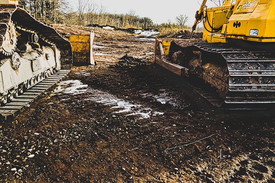 Two heavy machinery vehicles are parked on a muddy construction site, with one displaying a yellow color and the other in gray. The ground is uneven, featuring patches of exposed dirt and scattered gravel, with some snow visible in the background. Trees line the upper portion of the image against a cloudy sky.