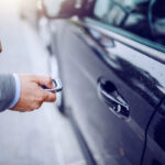 A man in a suit holds a car key remote near a black car's door, ready to unlock it.