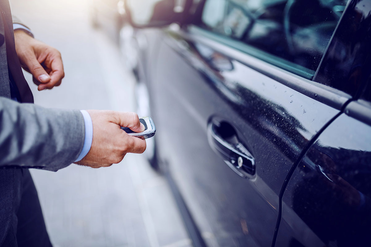 A man in a suit holds a car key remote near a black car's door, ready to unlock it.