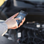 A person using a smartphone to access roadside assistance features while standing in front of an open car hood.