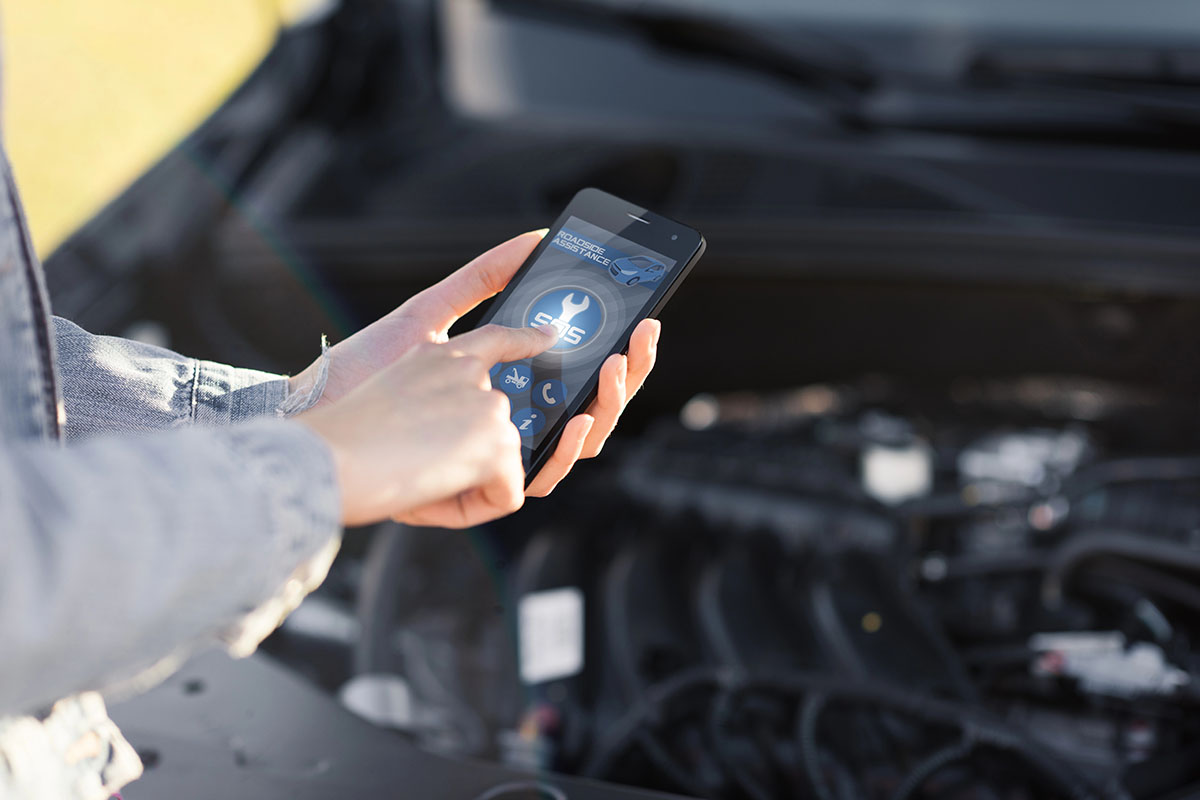 A person using a smartphone to access roadside assistance features while standing in front of an open car hood.