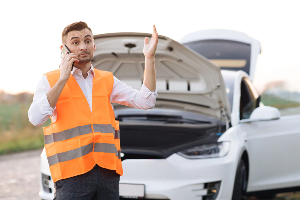 A man wearing an orange reflective vest stands beside a white car with its hood open, talking on a phone and gesturing with his hands in a roadside setting.