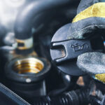 A close-up of a gloved hand holding an oil cap labeled with an oil can symbol, positioned above an engine, with visible hoses and components in the background.
