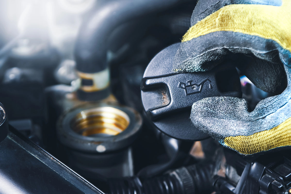 A close-up of a gloved hand holding an oil cap labeled with an oil can symbol, positioned above an engine, with visible hoses and components in the background.