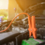 An orange jumper cable clamp attached to a car battery terminal inside an open engine bay.