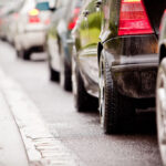 A low-angle view of bumper-to-bumper traffic lined up along a wet city curb, with red brake lights glowing.