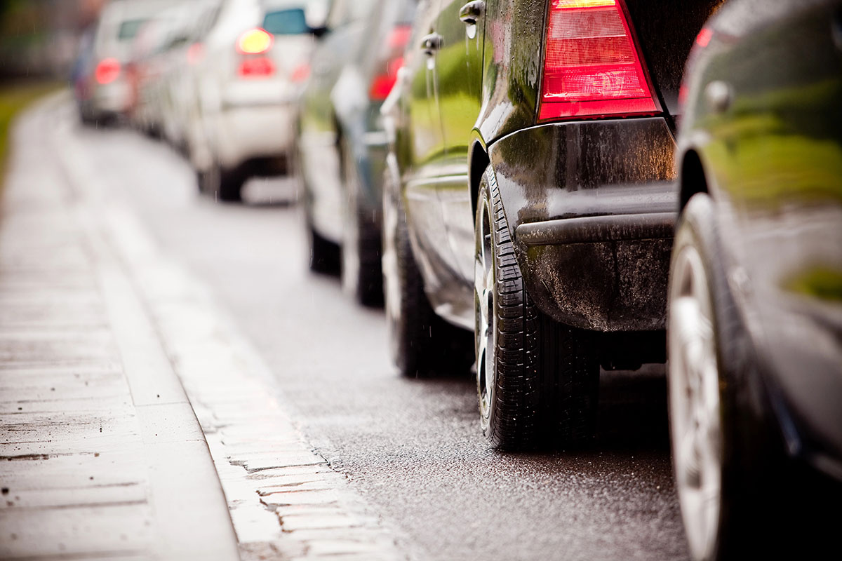 A low-angle view of bumper-to-bumper traffic lined up along a wet city curb, with red brake lights glowing.