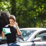 A young man leaning against a silver car with its hood popped open while holding a smartphone to his ear. He appears concerned as he looks down at a unfolded green paper document in his hand, with lush green trees in the background.