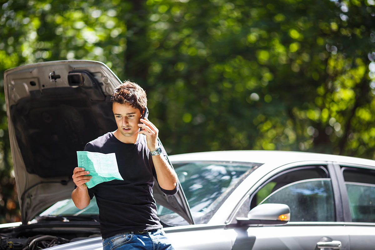 A young man leaning against a silver car with its hood popped open while holding a smartphone to his ear. He appears concerned as he looks down at a unfolded green paper document in his hand, with lush green trees in the background.