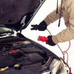 A person wearing black gloves and a winter jacket holding red and black jumper cable clamps near the open hood of a car engine bay, preparing to jump-start the vehicle outdoors in snowy winter conditions.