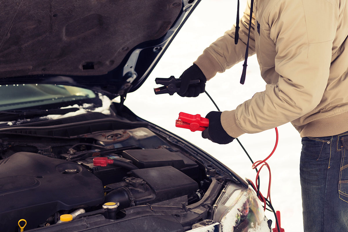 A person wearing black gloves and a winter jacket holding red and black jumper cable clamps near the open hood of a car engine bay, preparing to jump-start the vehicle outdoors in snowy winter conditions.