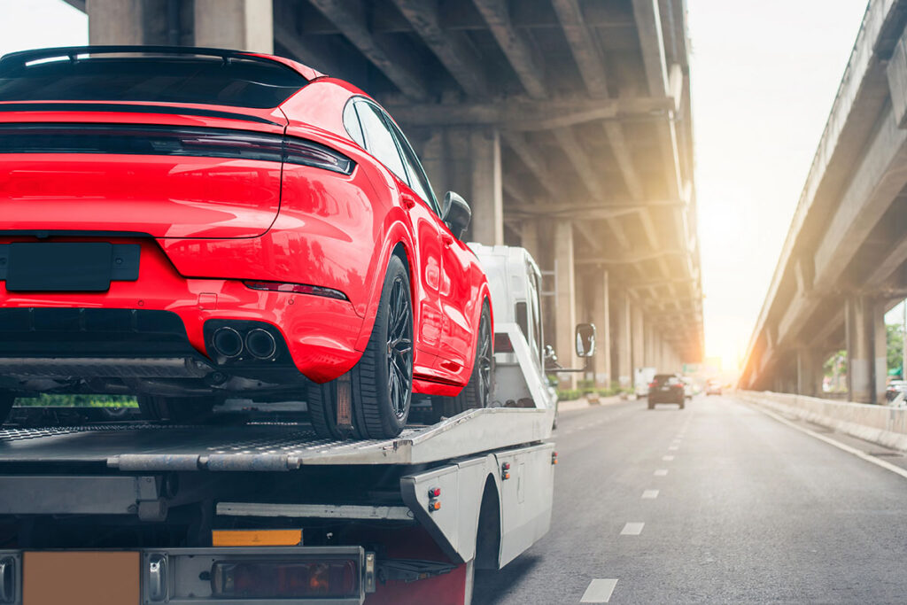 Low-angle rear view of a bright red sports SUV loaded onto the bed of a flatbed tow truck driving along a city highway.