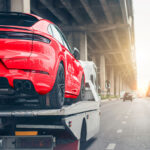 Low-angle rear view of a bright red sports SUV loaded onto the bed of a flatbed tow truck driving along a city highway.