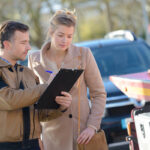 A male roadside assistance or tow truck worker in a brown jacket holding a clipboard while pointing to a document. A woman in a beige coat stands next to him, holding a pen and looking down at the clipboard, with a flatbed tow truck blurred in the background.