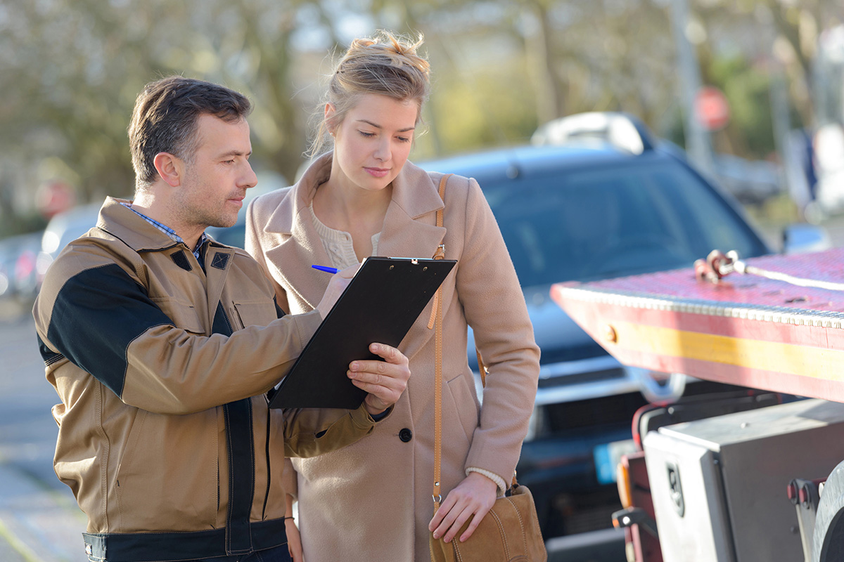 A male roadside assistance or tow truck worker in a brown jacket holding a clipboard while pointing to a document. A woman in a beige coat stands next to him, holding a pen and looking down at the clipboard, with a flatbed tow truck blurred in the background.